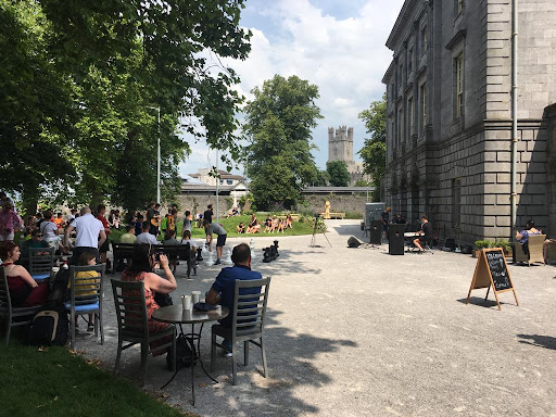 Outdoor café scene with people seated at tables, others gathering to watch a street performer. A historic building and distant tower are visible.