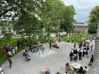 Outdoor scene with people gathered in a park. A band is performing while others watch a giant chess game. Groups chat at tables, creating a lively atmosphere.