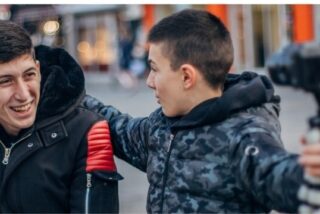 Two young men smiling, one with an arm around the other while holding a camera on a stick. They are outdoors, exuding a joyful, friendly vibe.