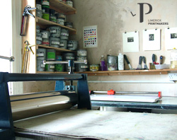 Studio interior with a printmaking press in the foreground. Shelves filled with colourful ink jars line the wall. Tools and prints hang nearby, creating a creative atmosphere.
