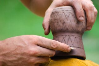 Hands meticulously carve intricate patterns into a clay pot on a yellow cloth backdrop, set against a blurred green background, conveying craftsmanship.