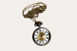 A vintage pocket watch with ornate hands and Roman numerals hangs from a long, intricate gold chain, evoking a sense of nostalgia and elegance.