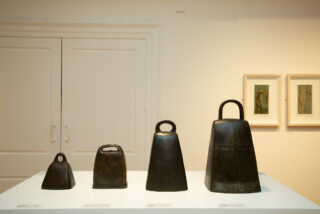Four cowbells of varying sizes displayed on a white pedestal, with two framed artworks in the background on a beige wall. The setting is calm and minimalistic.