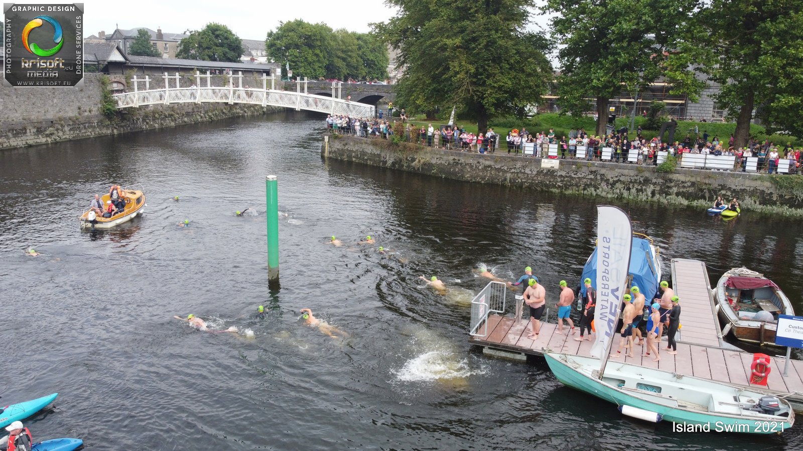 Aerial view of a river swim event with swimmers in green caps, a boat nearby, and onlookers on the riverbank. The atmosphere is lively.