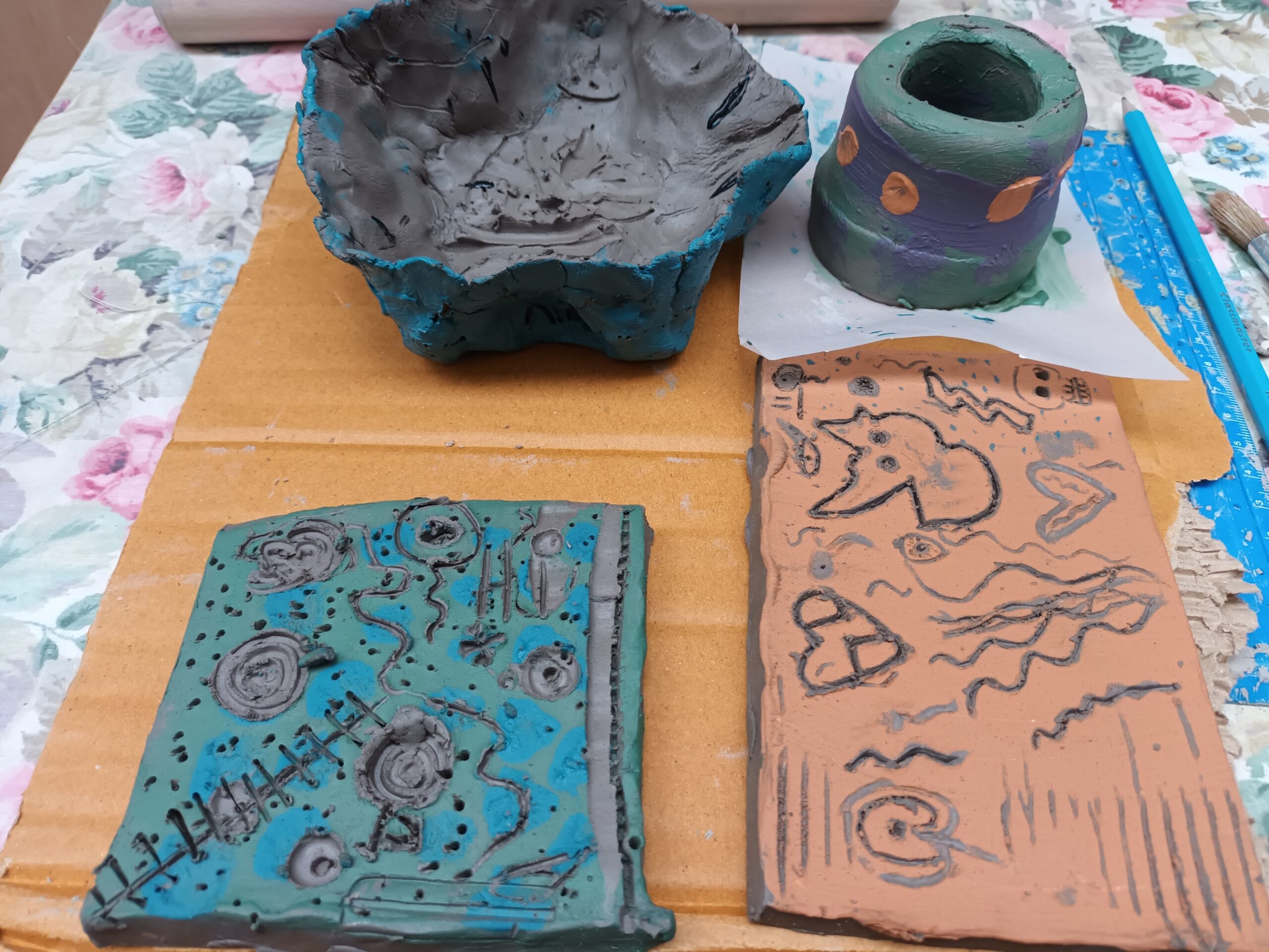 Colourful clay pottery and tiles on a floral tablecloth. A blue-grey bowl, a green-purple pot, and two carved tiles with abstract patterns evoke creativity.