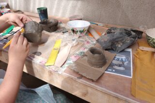 Hands shape clay on a wooden table filled with pottery tools, ceramic bowls, and fabric. The scene conveys creativity and focus in a pottery class.
