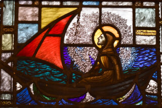 Stained glass image depicts a serene scene of a bearded man in a brown robe, with a halo, sailing on wavy waters under a large red sail.