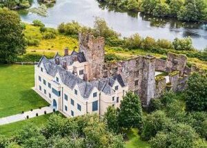 Aerial view of a historic castle with grey stone ruins and a well-preserved white building, surrounded by lush greenery and a serene river nearby.