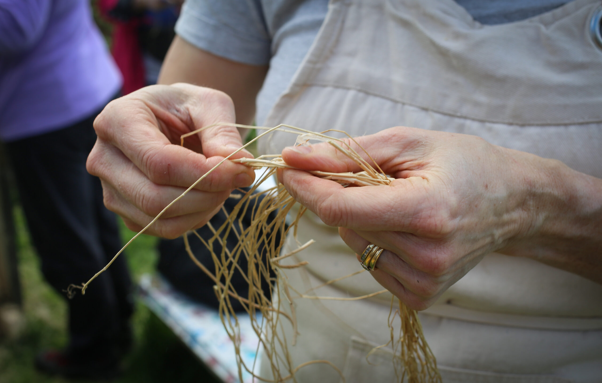 Hands weaving thin straw threads, creating a net-like pattern. The scene evokes craftsmanship and focus in an outdoor setting.