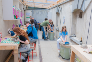 People in a sunny pottery studio work intently on projects, shaping clay on tables and wheels. The atmosphere is creative and focused.