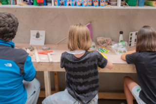 Three people sit at a cluttered workbench, engaged in crafts. Shelves above hold colourful paints. The atmosphere is creative and focused.