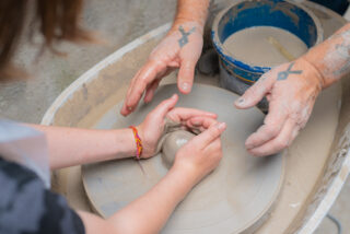 Close-up of hands at a potter's wheel; a child and an adult with tattoos shape wet clay together, symbolizing guidance and creativity.