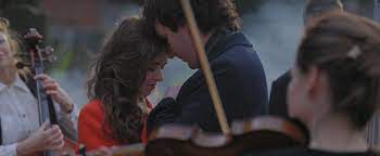 A couple shares an intimate moment, heads touching, surrounded by musicians with violins. They appear emotional and connected in a serene setting.