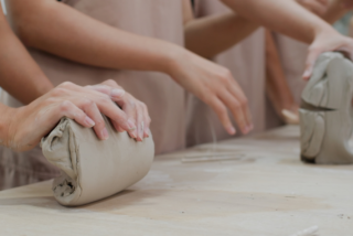 Hands of several people shaping clay on a wooden table, conveying focus and creativity in a pottery class. Aprons suggest an art studio setting.