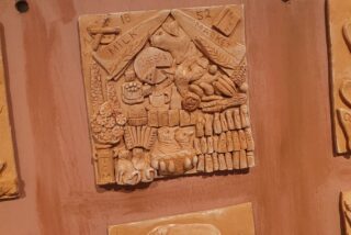 Clay relief sculpture depicting a bustling market scene, featuring items like milk, cheese, and baskets. Intricate details create a nostalgic, rustic feel.