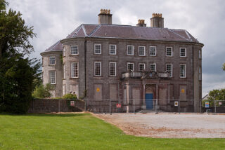 Historic stone mansion with a hipped roof and tall chimneys, surrounded by a metal fence. Overcast sky and lush greenery create a sombre tone.