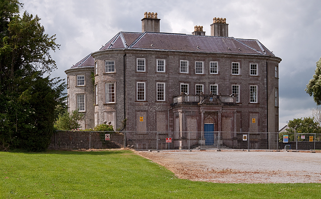 Historic stone mansion with a hipped roof and tall chimneys, surrounded by a metal fence. Overcast sky and lush greenery create a sombre tone.