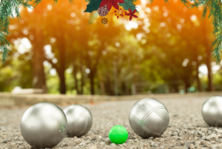 Silver bocce balls and a green target ball are on a gravel path, surrounded by trees. Festive pine and poinsettia decorations frame the scene.