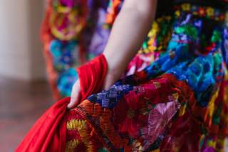 A person holds the edge of a vibrant, multi-coloured embroidered skirt, featuring intricate patterns and textures. The mood is lively and festive.