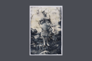 Faded black and white photo of a person in a coat, blurred face, holding a broom and basket, standing on rocky ground. The image has a vintage, ethereal feel.