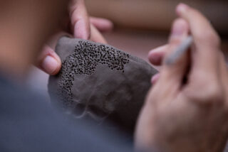 Hands meticulously carve intricate patterns of small holes into a clay surface, suggesting careful craftsmanship and artistic focus.