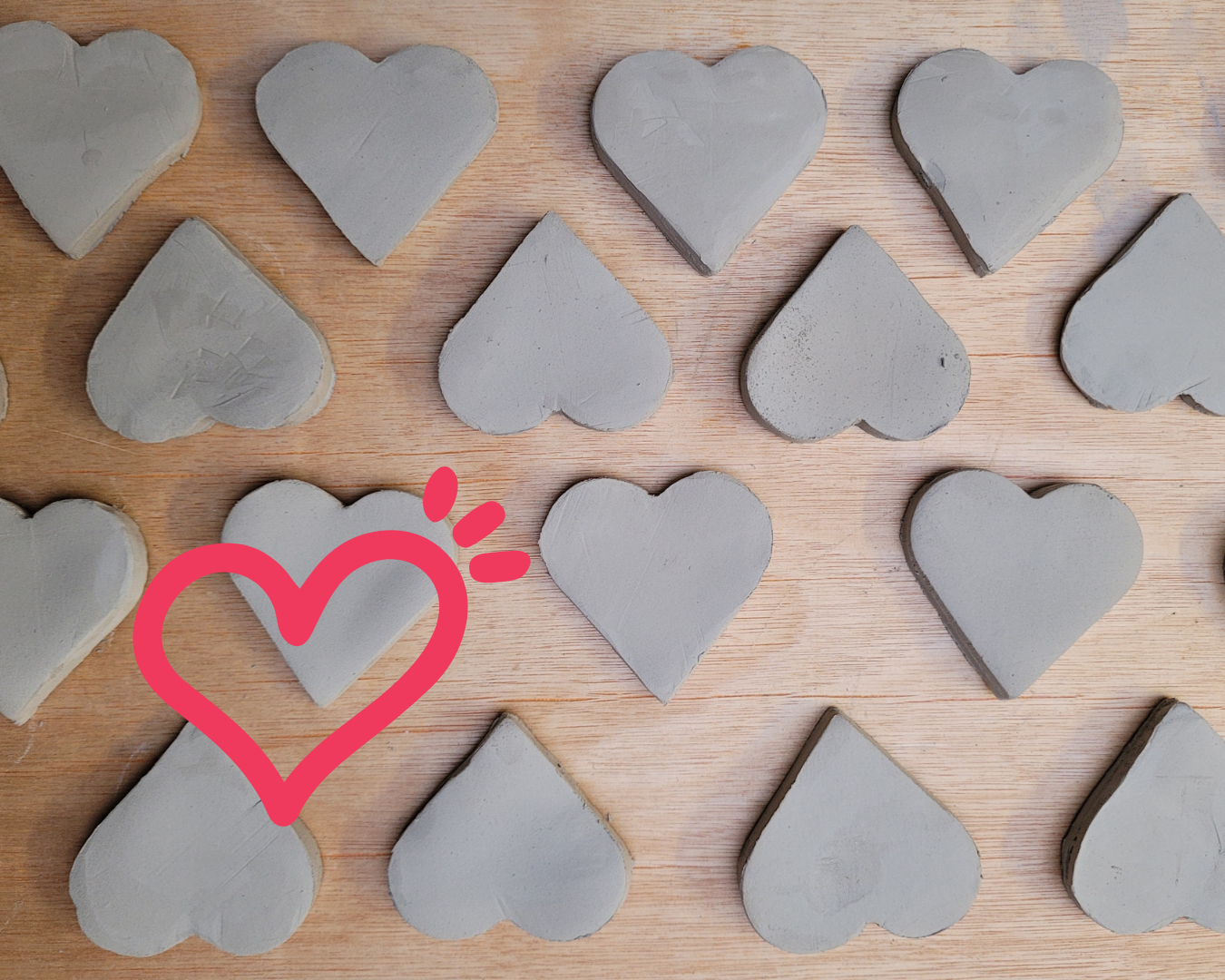 Concrete heart shapes laid on a wooden surface, evenly arranged. A pink heart sticker is in the lower left corner, adding a playful touch.