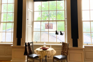 Cosy café setting in the Captain's Room with two chairs and a round table by a large window. The table has a yellow cloth, a dish, and a glass bottle. Soft daylight filters in, with tree and rooftop views outside. Two cat silhouettes and a bicycle sketch adorn the window, adding charm.
