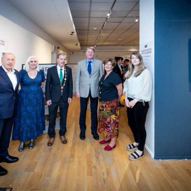 A group of six people stands in an art gallery. They are dressed in formal attire and appear to be part of an exhibition opening, exuding a celebratory tone. On the wall, there is a framed portrait and plaques with text, suggesting an educational or historical theme.