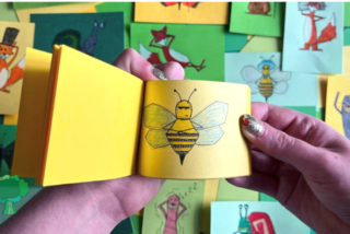 A hand holds a small yellow flipbook with a bee drawing. Background shows colourful paper cards with various cartoon animals, creating a playful, artistic scene.