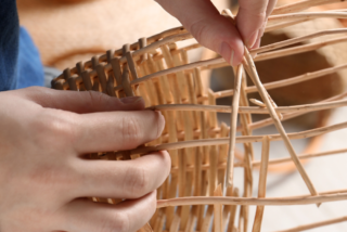 Hands weaving a wicker basket, with fingers skilfully threading thin strips. The setting is calm and focused, showcasing artisanal craftsmanship.