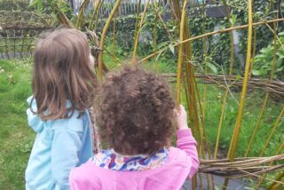 Two children in colourful outfits explore a garden structure made of woven branches, against a backdrop of a red-brick building and green grass.