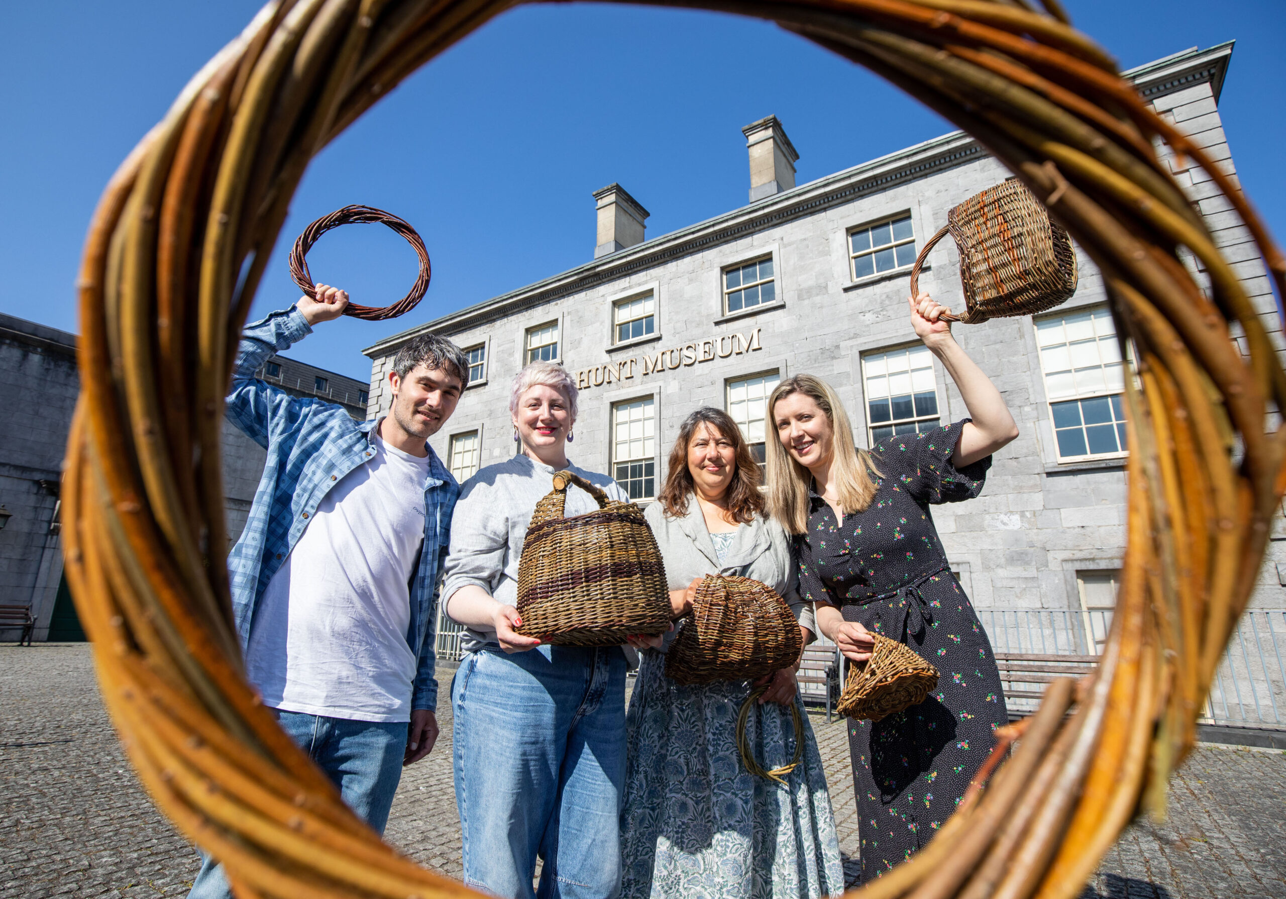 Four people proudly hold woven baskets in front of the Hunt Museum. Framed by a large wicker hoop, the scene conveys creativity and heritage under a clear blue sky.