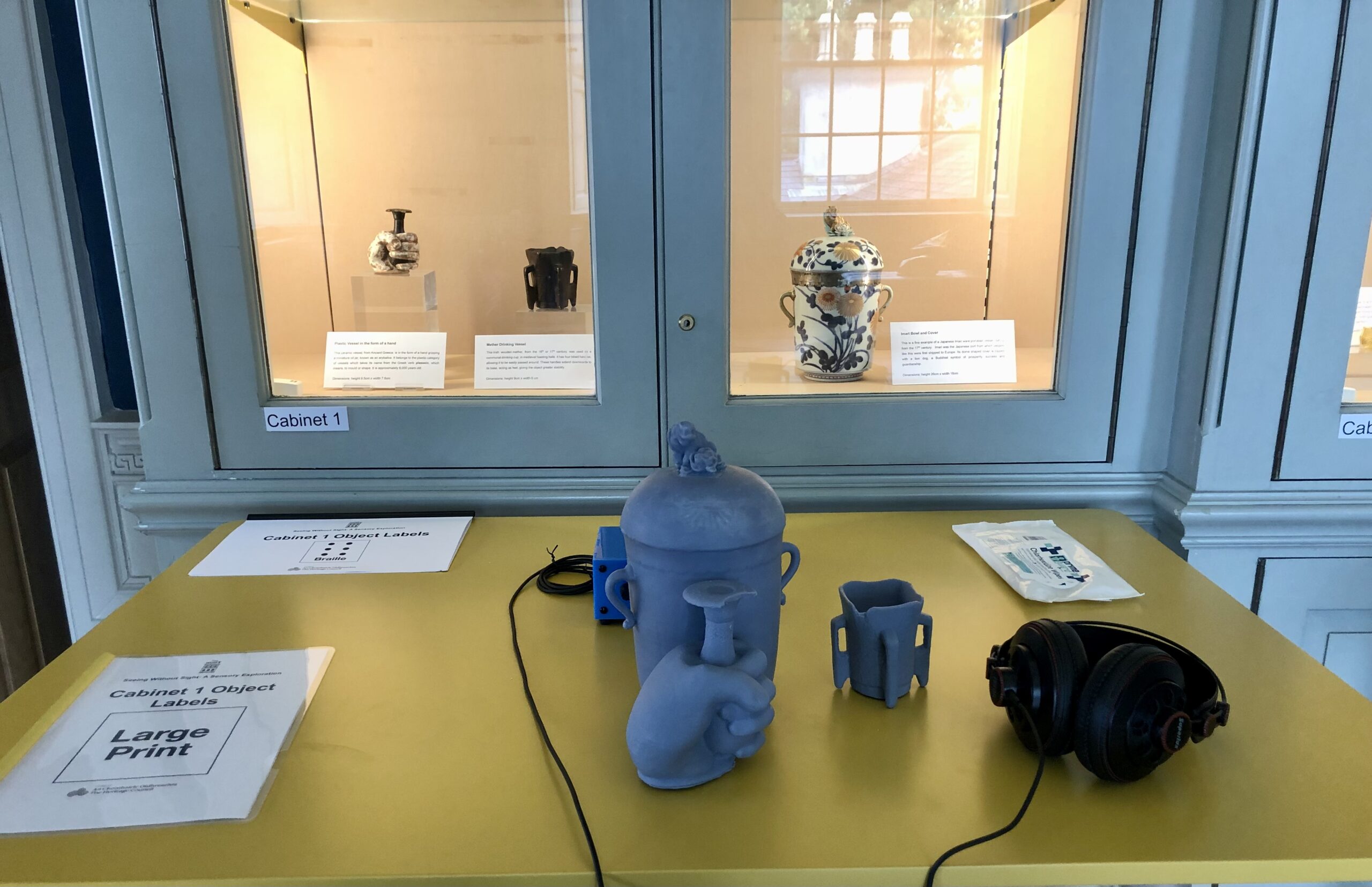 Museum display features ceramics in glass cabinet and tactile replicas on a yellow table, with large-print labels and headphones for audio descriptions.