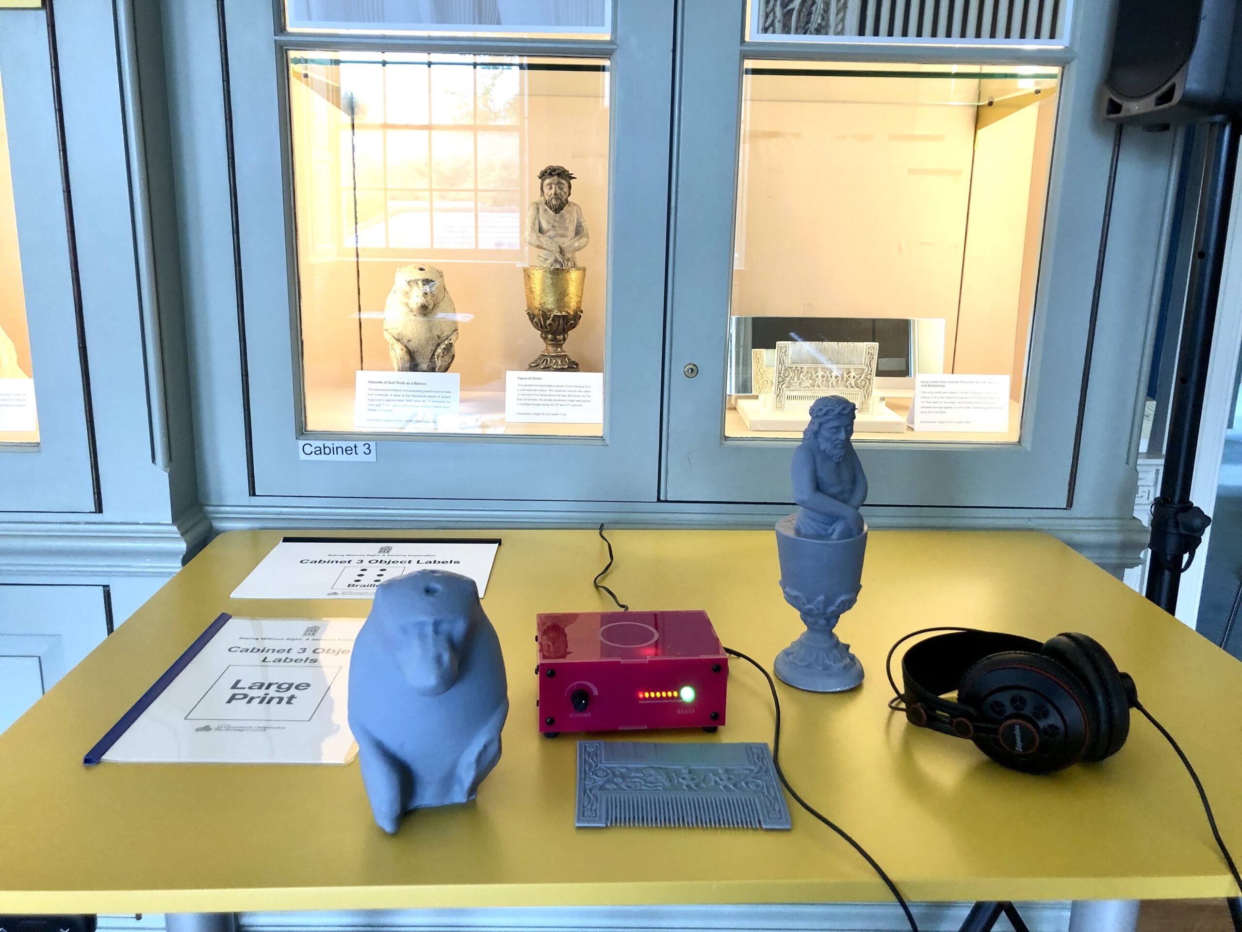 A yellow table holds 3D-printed statues, headphones, and a red audio device. Behind, museum artifacts are displayed in a glass cabinet labelled 