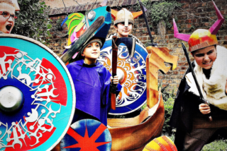 Children in colourful Viking costumes playfully pose with shields and swords. The scene is lively, set against a rustic brick wall and greenery.