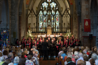 A choir, wearing black with pink scarves, performs in a church with a large stained glass window. An audience fills the pews, creating a serene atmosphere.