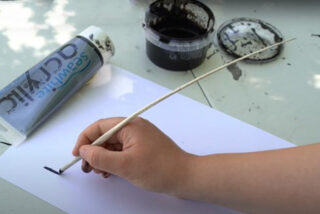 A hand paints with black acrylic on white paper using a brush. Nearby are a tube of paint and an open container of black paint, conveying creativity.