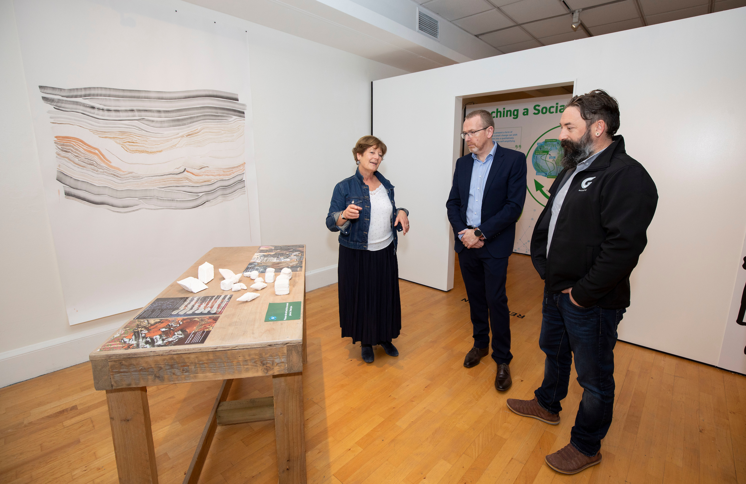 Three people stand in a gallery. A woman gestures while explaining. A large abstract artwork and a wooden table with objects are visible.