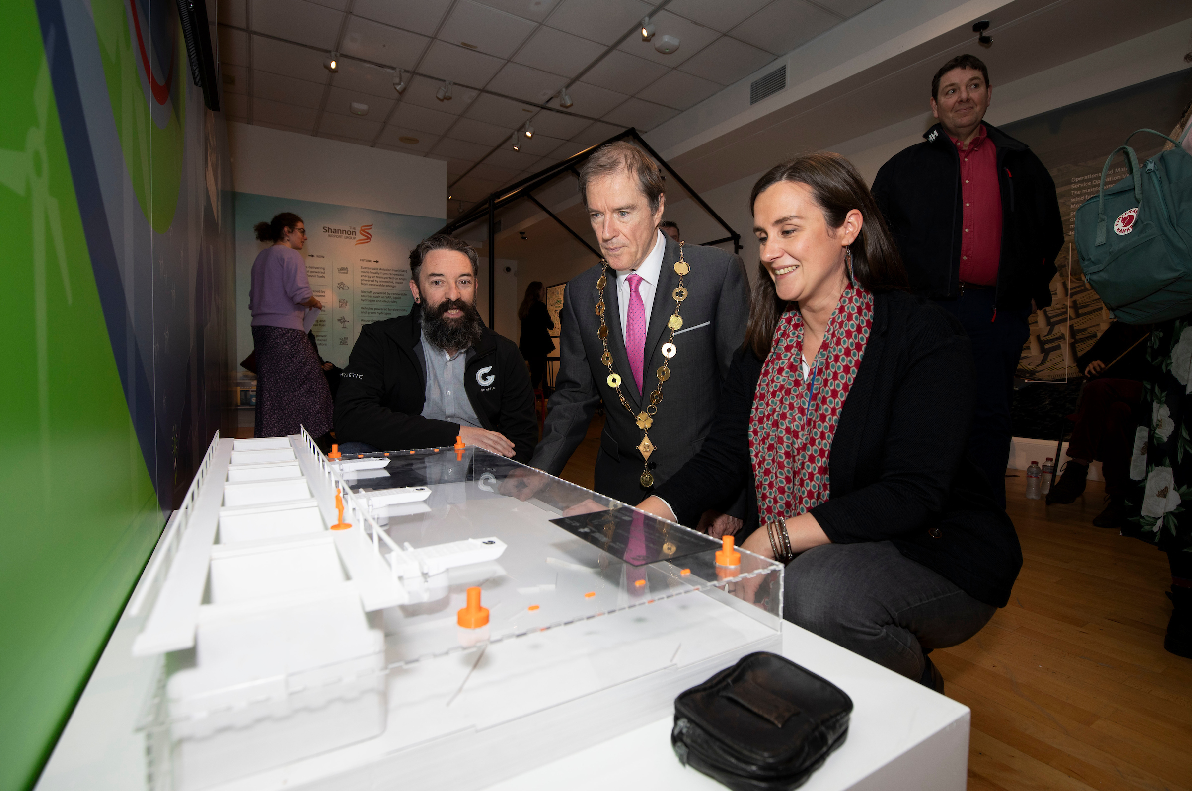A man in a suit with a chain of office examines a scale model on a table alongside two people. The setting is a brightly lit exhibition space.