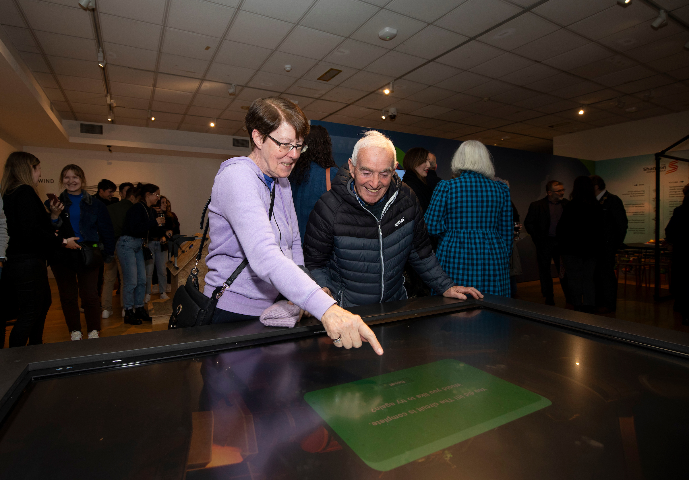 A woman and an older man enthusiastically interact with a large touchscreen in a museum, surrounded by a small crowd. The atmosphere is lively and engaging.