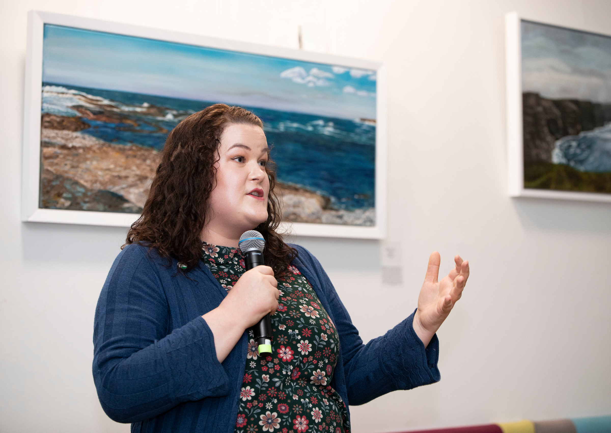A woman with curly hair speaks into a microphone in an art gallery, gesturing expressively. Coastal landscape paintings are displayed on the wall behind her.
