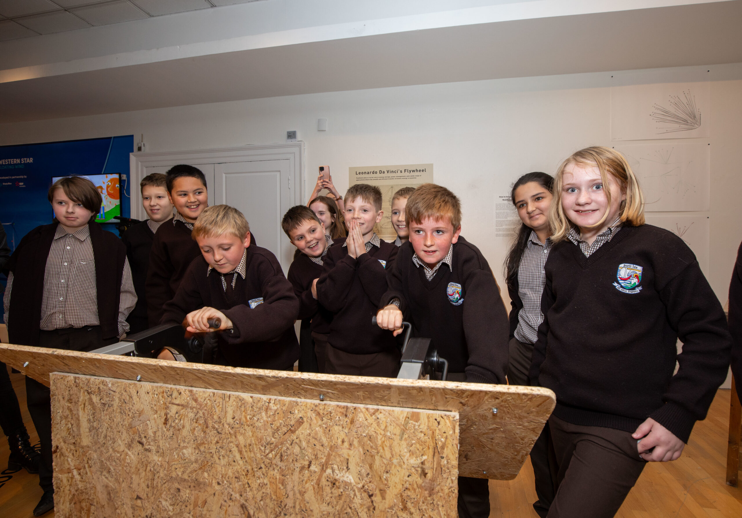 A group of schoolchildren in uniform enthusiastically gather around a wooden mechanical exhibit. The room has educational displays and a lively atmosphere.