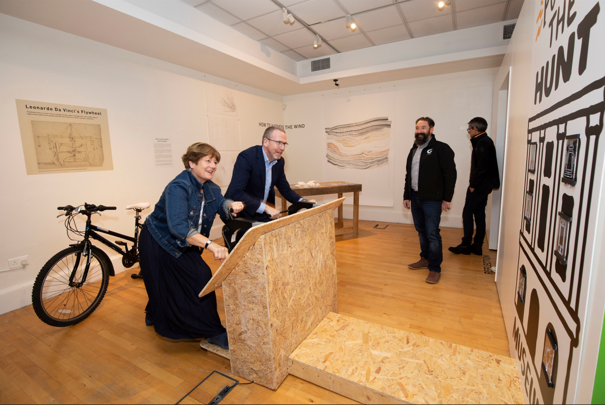 Two smiling people engage with an interactive exhibit in a gallery featuring Leonardo da Vinci's flywheel and other artworks. Bicycles and observers are present.