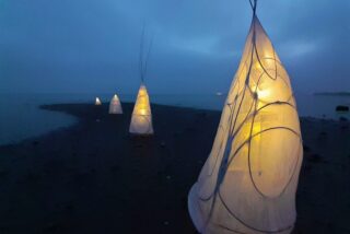 Glowing fabric tepees on a dark, misty beach. The structures, softly lit from within, create a serene, mystical ambiance along the shoreline.