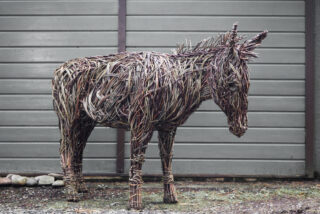 A life-sized donkey sculpture made of woven twigs stands in front of a grey wooden wall. The intricate weaving conveys a rustic, earthy feel.