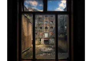 View through a dark window of a dilapidated brick building with many small windows. Sky is partly cloudy. Mood: abandoned, sombre, quiet.
