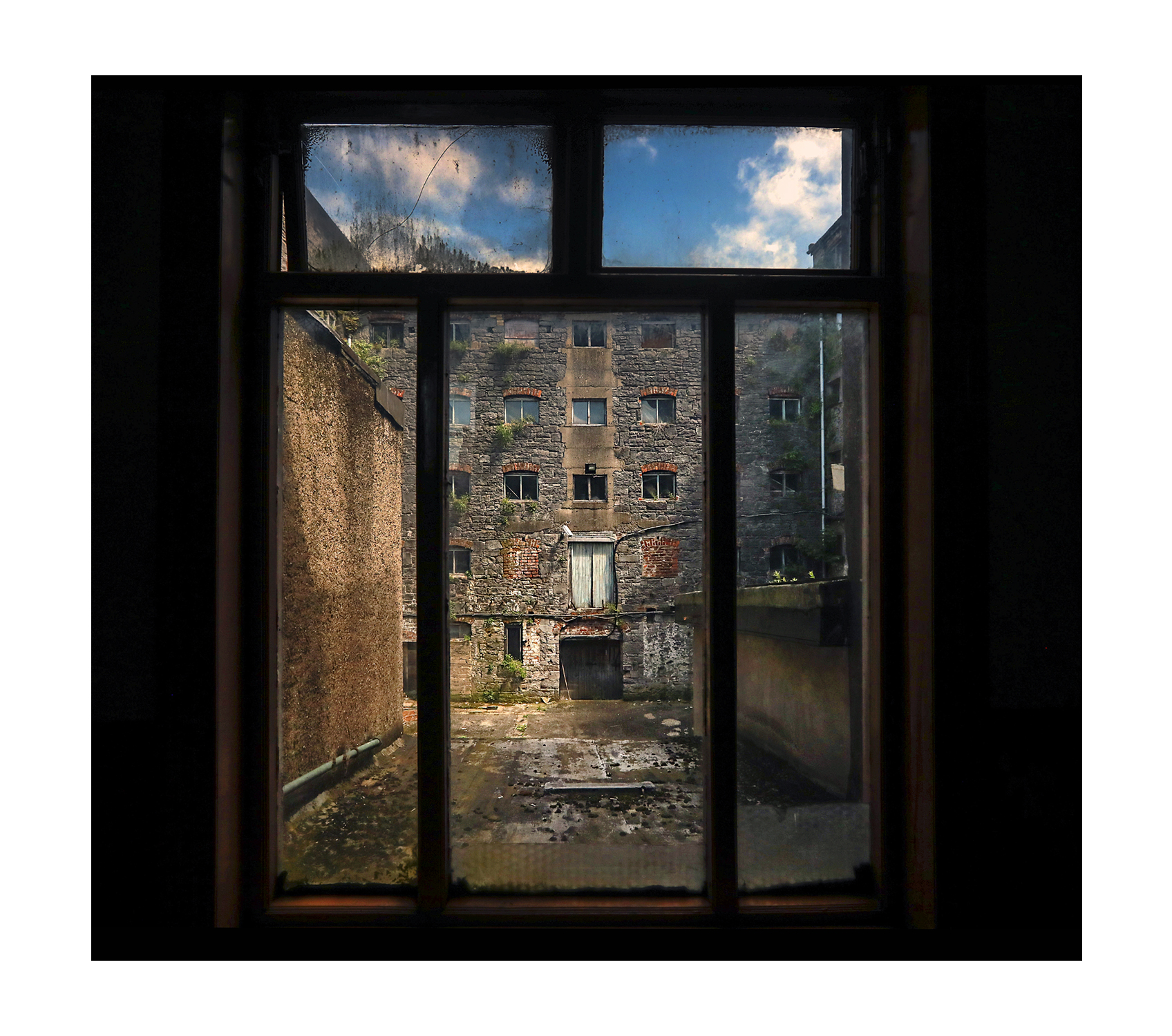View through a dark window of a dilapidated brick building with many small windows. Sky is partly cloudy. Mood: abandoned, sombre, quiet.