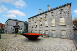 The image shows the exterior of The Hunt Museum, a historic stone building with large windows. In front, there is a modern red metal sculpture on a cobblestone courtyard. The sky is clear and blue, giving a serene and inviting atmosphere.