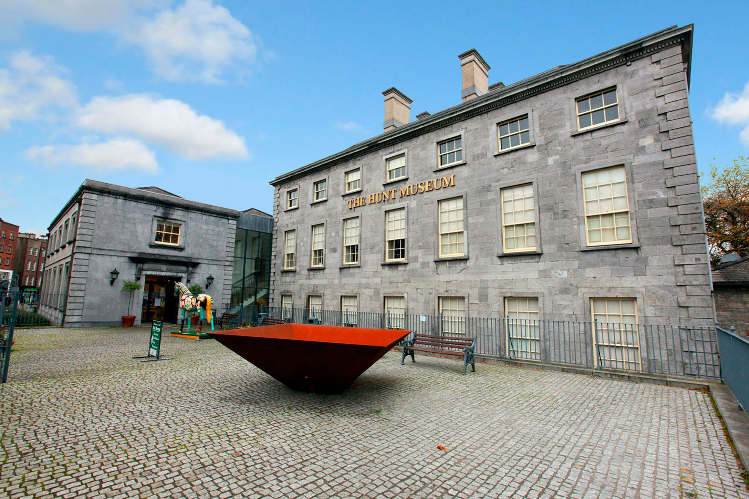 The image shows the exterior of The Hunt Museum, a historic stone building with large windows. In front, there is a modern red metal sculpture on a cobblestone courtyard. The sky is clear and blue, giving a serene and inviting atmosphere.