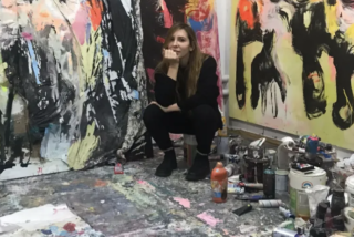 A person sits contemplatively in a vibrant, paint-splattered studio surrounded by abstract, colourful paintings, conveying a creative and chaotic atmosphere.